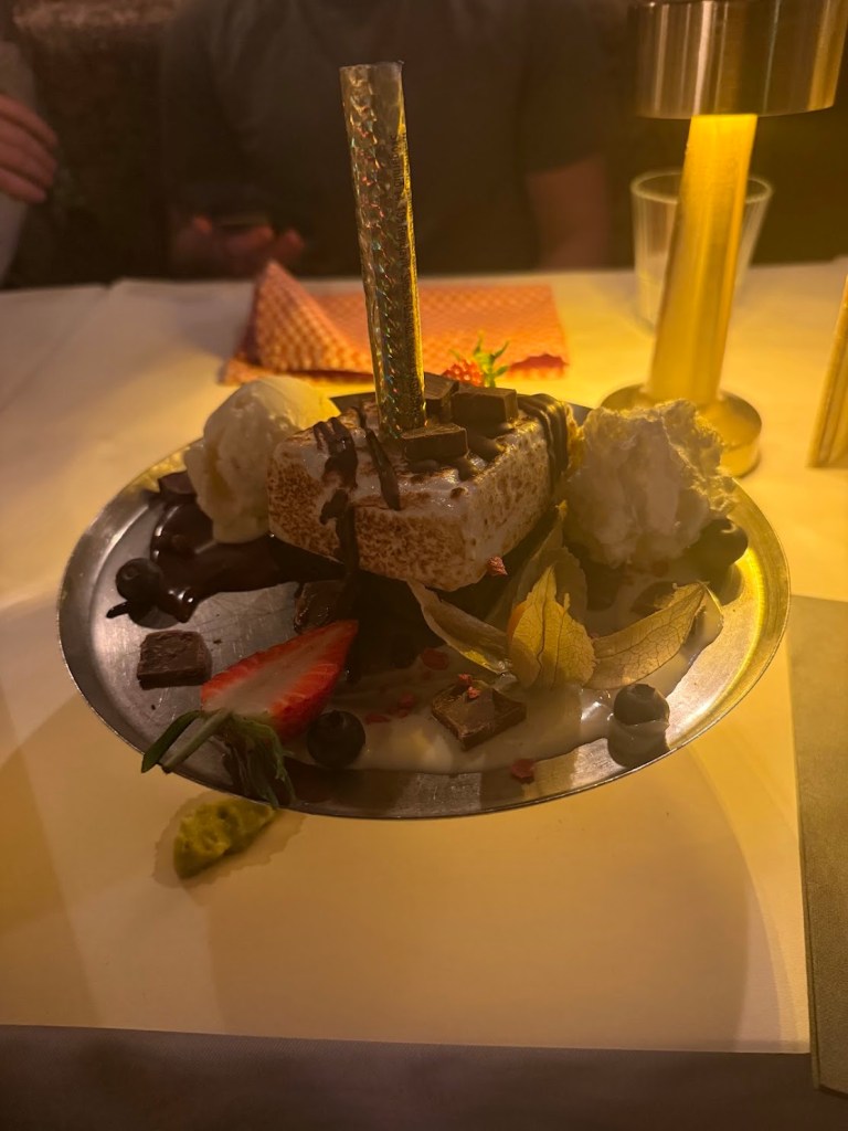 A plate of dessert featuring a piece of cake with chocolate drizzle, topped with whipped cream, strawberries, blueberries, and decorative leaves, accompanied by a shiny, colorful straw.