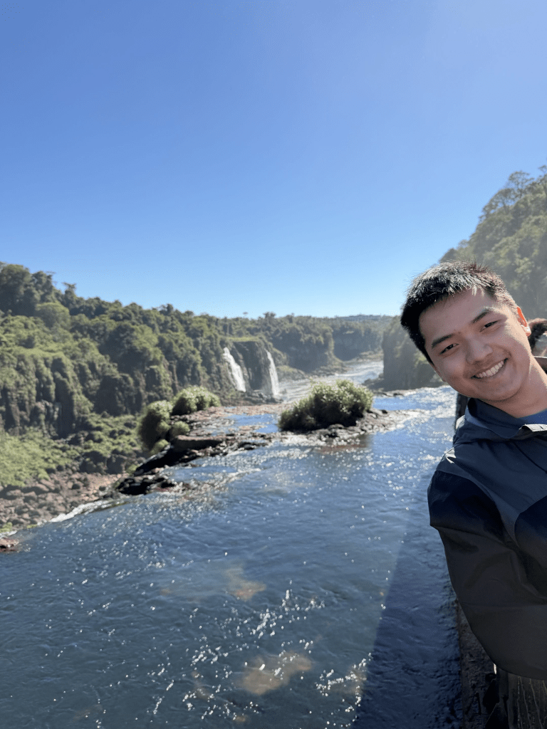 A smiling individual posing on a bridge near a scenic waterfall and river surrounded by lush greenery and clear blue sky.