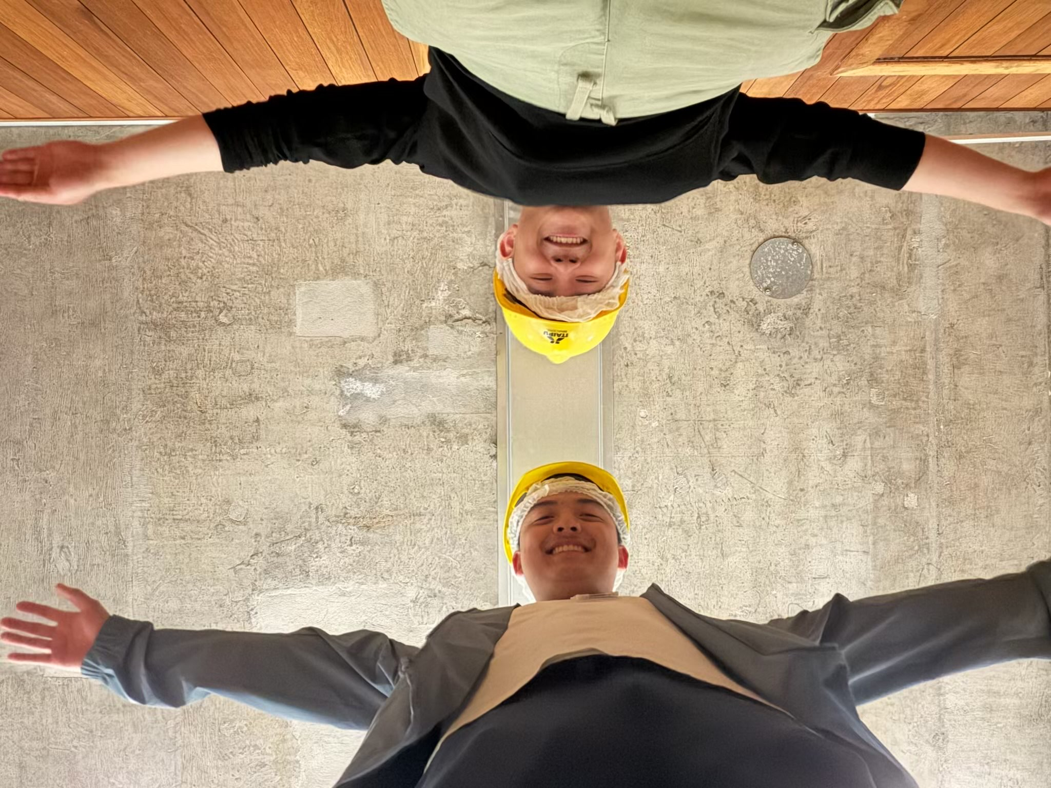 Two smiling individuals, wearing yellow hard hats, are standing with their arms outstretched. The photo is taken from below, showing them from feet to face against a textured background.
