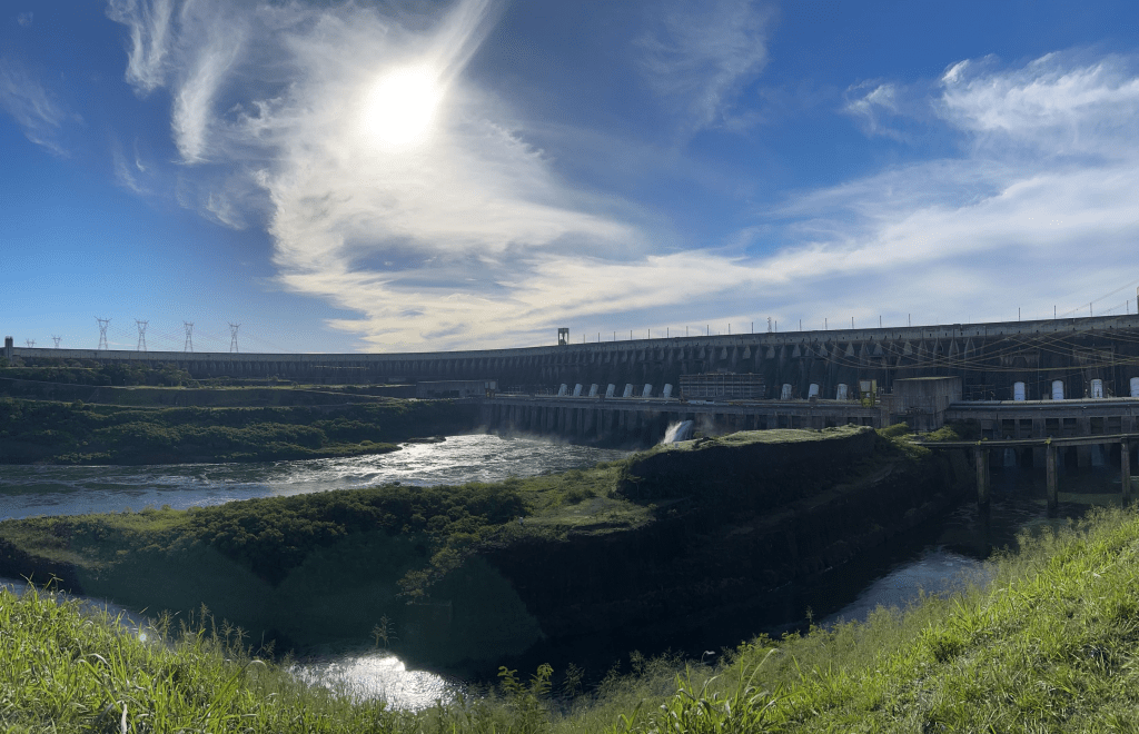 A panoramic view of a hydroelectric dam with a river in the foreground, surrounded by lush greenery and a clear blue sky with fluffy clouds.